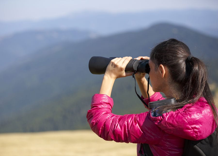 A woman looking through binoculars while standing on top of a hill.
