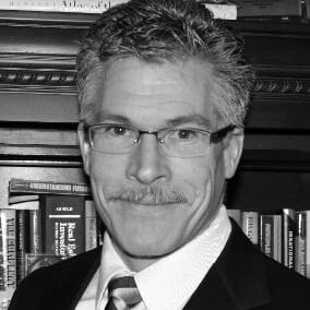 A man with glasses and a mustache in front of a bookshelf.